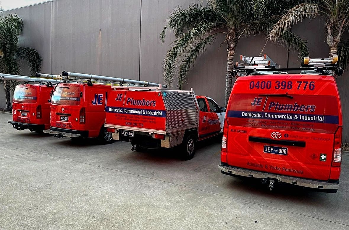 The JE Plumbers fleet of red branded vans and ute lined up, signwritten with Domestic, Commercial and Industrial and the phone number 0400 398 776