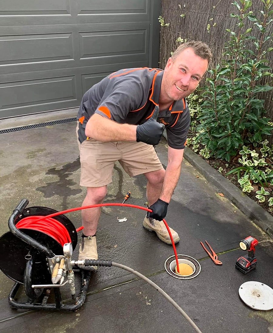 Jake from JE Plumbers clearing a blocked drain with a drain machine on a driveway, in branded uniform