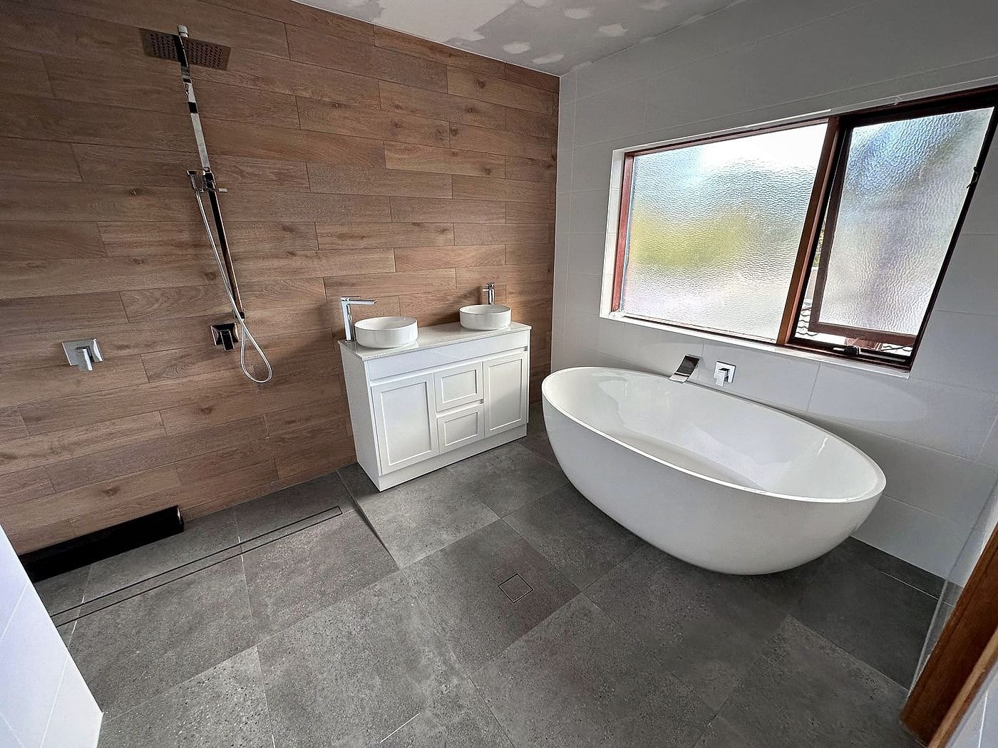 Modern bathroom renovation by JE Plumbers with a freestanding bath, timber feature wall, twin vessel basins and a chrome rainfall shower